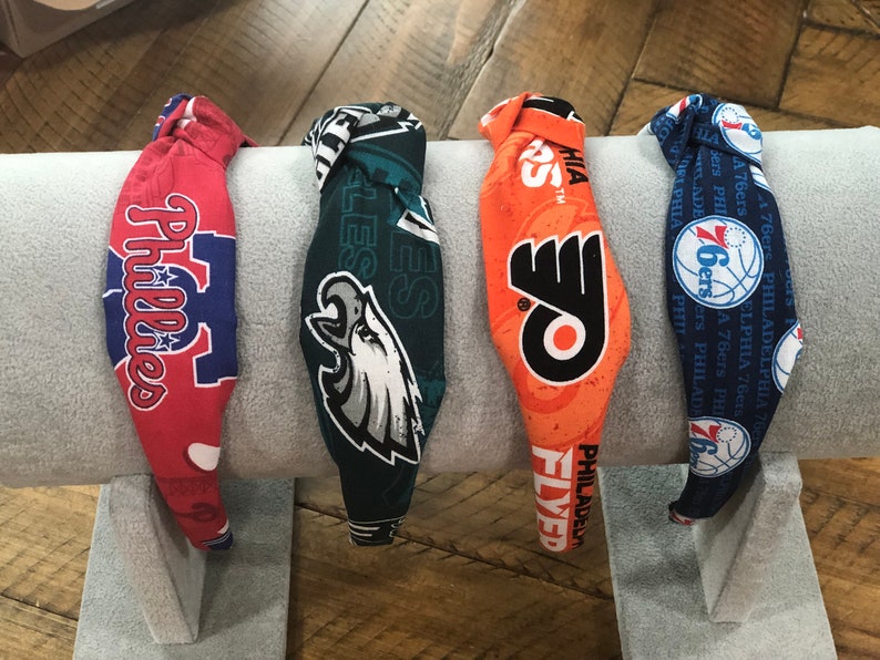 May include: Four fabric headbands featuring logos of Philadelphia sports teams: Phillies, Eagles, Flyers, and 76ers. The headbands are red, green, orange, and blue, with team logos and text. They are displayed on a gray, padded surface.
