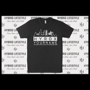 May include: Dark gray t-shirt with a white skyline graphic and the text "HYROX YOURNAME HOUSTON 2026." The shirt also features the text "HYBRID LIFESTYLE Train Hard. Live Bold." repeated on the background.
