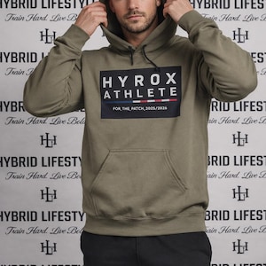 May include: An olive green hoodie with a front pocket and a hood. The front of the hoodie features a black rectangle with the text "HYROX ATHLETE" and "FOR, THE PATCH, 2025/2026". The background has the words "HYBRID LIFESTYLE" repeated.