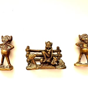 Könnte beinhalten: Drei Messingfiguren von skurrilen Charakteren. Zwei Figuren stehen mit den Händen in den Hüften, während die dritte auf einem Zaun mit einem Pilz sitzt. Die Figuren haben ein Vintage- und antikes Aussehen.