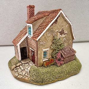 May include: A detailed miniature cottage figurine with a red-tiled roof and brick chimney. The cottage has beige walls, a small door, and windows. It sits on a green base with a stone pathway and miniature shrubs.