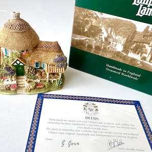 Könnte beinhalten: Eine detaillierte Miniatur eines Cottage mit Strohdach, grüner Tür und farbenfrohen Blumen- und Fruchtdetails. Das Cottage wird von einer Schachtel und einer Eigentumsurkunde begleitet. Auf der Schachtel steht "Handmade in England, Treasured Worldwide".