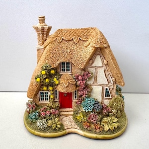 Può includere: Figurina di una casa di campagna in miniatura con tetto di paglia, porta rossa e dettagliato paesaggio floreale. La casa presenta pareti bianche con struttura in legno marrone e piccole finestre. Il camino è marrone e la base è verde chiaro con fiori colorati.