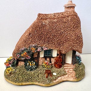 Könnte beinhalten: Miniatur-Figur eines Cottage mit Strohdach und Schornstein. Das Cottage ist mit bunten Blumen und Grün geschmückt. Eine Geige steht neben der Tür. Die Figur besteht aus einem strukturierten Material.