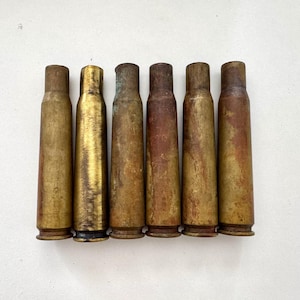 May include: Six antique brass bullet casings, displayed in a row. The casings range in colour from bright gold to a weathered brown, indicating age and use. Each casing is cylindrical with a flared base.
