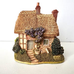 Puede incluir: Figurita en miniatura de una casa de campo con tejado de paja y chimenea de ladrillo. Las paredes de piedra presentan una pequeña puerta, una ventana y escalones. Flores moradas y follaje verde adornan la casa. Una pieza decorativa encantadora.