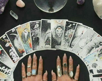 May include: A tarot card reading spread with a crystal ball and crystals on a black surface. The cards are fanned out in a semi-circle with the Major Arcana cards visible. The cards include The World, Strength, The Hanged Man, and The Tower. The hands of a person are visible in the centre of the spread, wearing rings and nail polish.