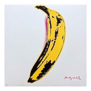 May include: A square art print featuring a stylized yellow banana with black textured details and a pink shadow outline against a white background. The signature Andy Warhol is printed in red in the bottom right corner with a small stamp in the bottom left.