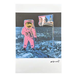 Puede incluir: Estampado de arte pop que presenta un astronauta rosa sobre una superficie lunar azul y negra con una bandera estadounidense. El casco del astronauta tiene una visera amarilla. La obra est&aacute; firmada en la esquina inferior derecha.