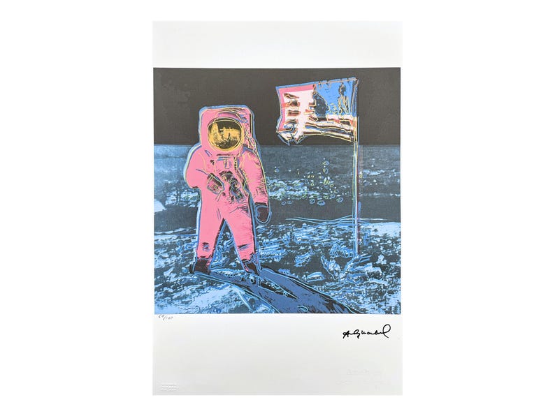 Puede incluir: Estampado de arte pop que presenta un astronauta rosa sobre una superficie lunar azul y negra con una bandera estadounidense. El casco del astronauta tiene una visera amarilla. La obra est&aacute; firmada en la esquina inferior derecha.