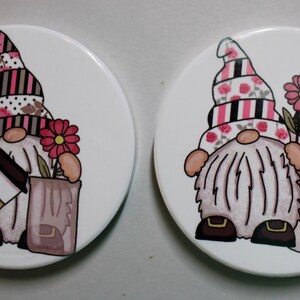 May include: Two white ceramic coasters with a gnome design. Each gnome is wearing a pink and black striped hat with floral accents. One gnome is holding a coffee cup and the other is holding a bag with flowers.