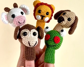 May include: A set of five crocheted finger puppets featuring animal heads including a white cow, a yellow lion with a brown mane, a tan dog, a brown monkey, and a green frog with large white eyes and red circular pupils, all with black safety eyes.