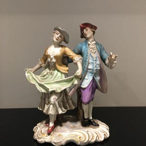 May include: A porcelain figurine of a couple dancing. The woman is wearing a green and white dress with a yellow jacket and a grey hat. The man is wearing a blue jacket with a purple waistcoat and a red hat. The figurine is on a white base with gold accents.