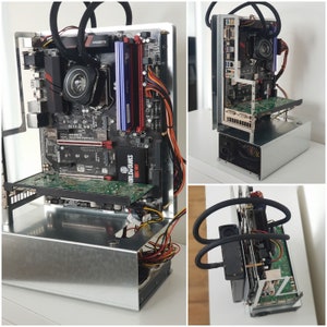 Könnte beinhalten: Ein selbstgebauter Computer mit einem silbernen Gehäuse, einem Motherboard mit einem schwarzen und roten Kühlkörper und einer transparenten Acrylscheibe. Der Computer ist mit einem schwarzen und silbernen Netzteil und einer schwarzen und roten Grafikkarte verbunden. Der Computer steht auf einer weißen Oberfläche.