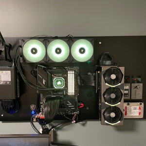 Op de afbeelding: Een zwart computer moederbord met drie witte ventilatoren en een zwarte grafische kaart met drie witte ventilatoren. Het moederbord en de grafische kaart zijn gemonteerd op een zwarte muur.