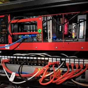 Peut inclure: Un boîtier d'ordinateur en métal rouge avec une carte mère noire et de nombreux câbles. Le boîtier est monté dans un rack avec un commutateur réseau noir en dessous. Le commutateur a 16 ports et est connecté à plusieurs câbles rouges et blancs.