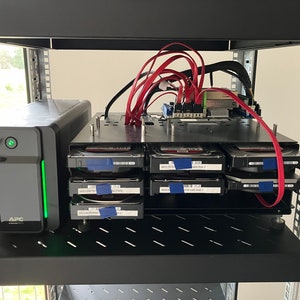 Könnte beinhalten: Ein schwarzer Serverrack mit einem schwarzen USV-Gerät und mehreren Festplatten. Die Festplatten sind mit blauem Klebeband beschriftet und der Serverrack ist mit roten Kabeln an eine Stromquelle angeschlossen.