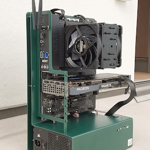 Puede incluir: Una caja de ordenador de metal verde con una placa base negra, una tarjeta gráfica negra con el texto "GIGABYTE" y "RADEON", y una fuente de alimentación negra. La caja tiene dos antenas en la parte posterior.