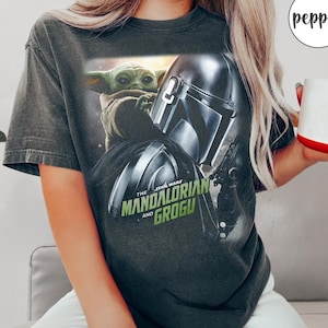 May include: Dark gray t-shirt featuring a graphic of Grogu and the Mandalorian helmet from The Star Wars series. The text "The Mandalorian and Grogu" is printed in green and white. The shirt has short sleeves and a crew neck.