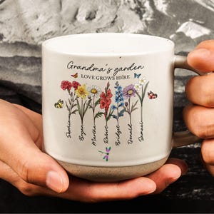 May include: White ceramic mug with a floral design and the text "Grandma's garden LOVE GROWS HERE". The mug has a brown rim and base. The floral design includes a variety of flowers and butterflies. The names Sophia, Bryen, Martha, Robert, Bridget, Donald, and Samuel are written below the flowers.
