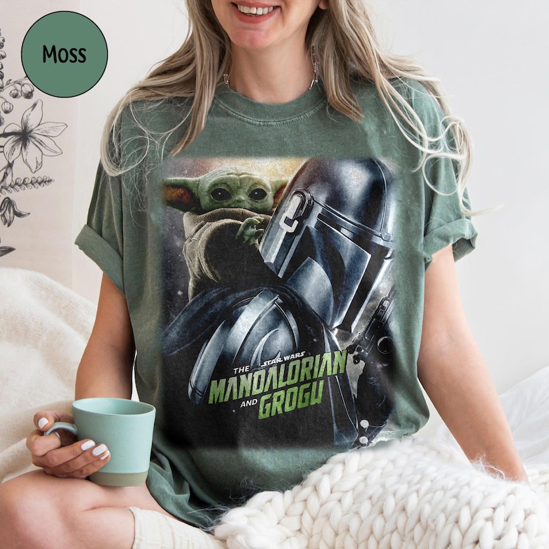 May include: Moss green t-shirt featuring a graphic of The Mandalorian and Grogu from Star Wars. The design includes the Mandalorian's helmet and Grogu's face. The text "THE STAR WARS" and "MANDALORIAN AND GROGU" are printed on the shirt.