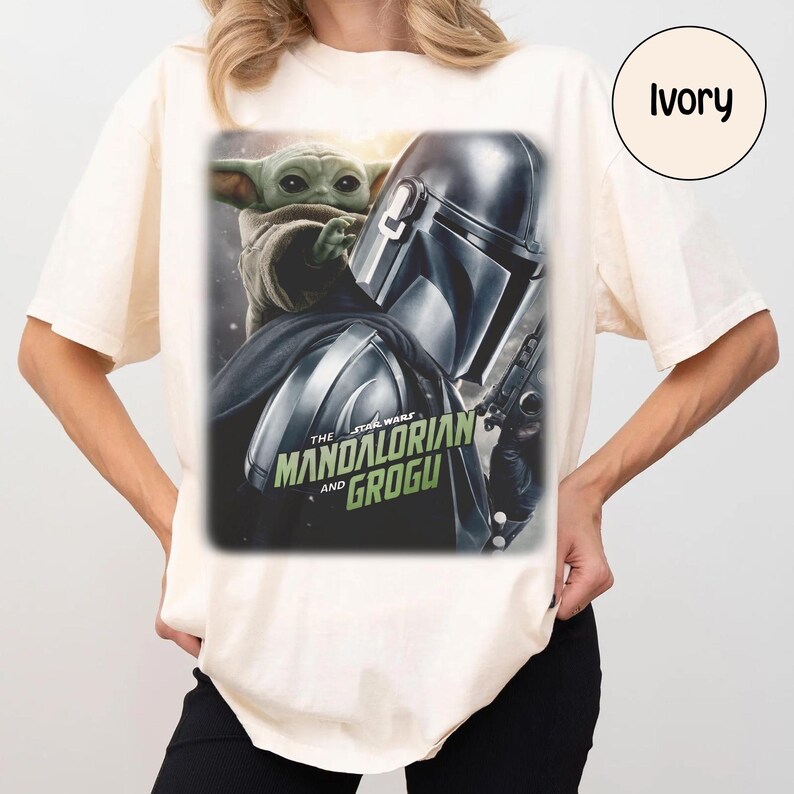 May include: Ivory-colored t-shirt featuring a graphic from The Mandalorian series. The design showcases Grogu and the Mandalorian, with the text "The Mandalorian and Grogu" in green and white. The shirt is a casual wear item.