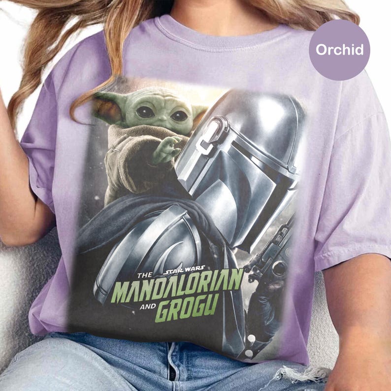 May include: Orchid-colored t-shirt featuring a graphic of Grogu and The Mandalorian from Star Wars. The design includes Grogu, the Mandalorian's helmet, and the text "The Mandalorian and Grogu".