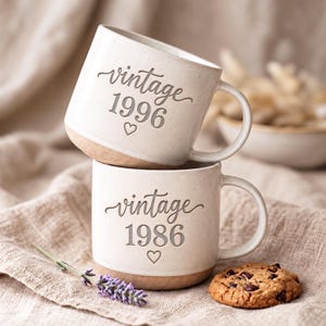 May include: Two off-white ceramic mugs stacked vertically. The top mug features the text vintage 1996 with a small heart below. The bottom mug features the text vintage 1986 with a small heart below. Both mugs have a speckled finish and a handle.