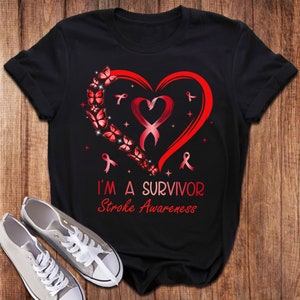 Peut inclure: T-shirt noir avec un motif de cœur rouge orné de papillons et de rubans rouges. Le texte "I'm a Survivor Stroke Awareness" est imprimé sous le cœur.