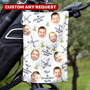 May include: White golf towel with a repeating pattern of golf balls, clubs, and smiling faces. The text "Best Grandpa By Par" is also printed on the towel. The towel is hanging on a golf cart.