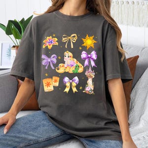 May include: A dark gray t-shirt with a graphic print of Rapunzel from the Disney movie Tangled. The graphic includes Rapunzel, a tower, a sun, flowers, and lanterns.