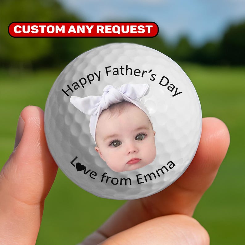May include: A white golf ball with a baby's face and a white bow headband. The text reads "Happy Father's Day Love from Emma". The top of the ball has the text "CUSTOM ANY REQUEST" in a red banner.