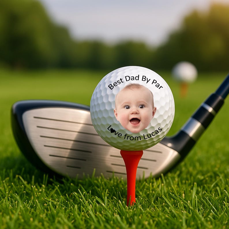 May include: A personalized golf ball with a baby's face, the text "Best Dad By Par" and "Love from Lucas". The ball is on a red tee, with a golf club in the background, set on a green golf course.