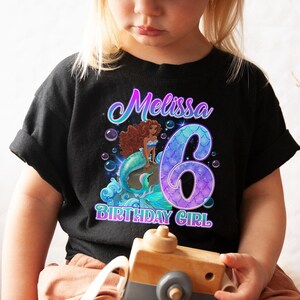 Puede incluir: Camiseta negra con un gr&aacute;fico de sirena y el texto "Melissa 6 Birthday Girl". La sirena est&aacute; sentada en una roca con una cola iridiscente p&uacute;rpura y azul.