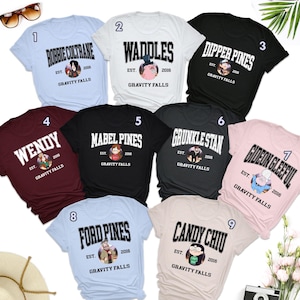 May include: Nine different t-shirts featuring characters and text from the animated television series Gravity Falls. The shirts are in various colors, including light blue, white, black, maroon, pink, and gray. Each shirt has a different character or text design, including Robbie, Waddles, Dipper Pines, Wendy, Mabel Pines, Grunkle Stan, Gideon Gleeful, Ford Pines, and Candy Chiu.