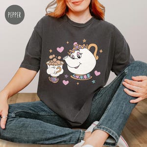 May include: Dark grey t-shirt with a cartoon teapot and teacup design, pink hearts and stars. The teapot has a smiling face and a crown. The shirt is a comfortable, casual style, suitable for everyday wear.