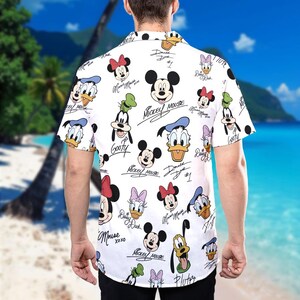 May include: White short-sleeved button-down shirt with a colourful all-over print of Disney characters, including Mickey Mouse, Minnie Mouse, Donald Duck, Goofy, and Pluto. The characters are depicted in a cartoon style with their signature names and autographs.