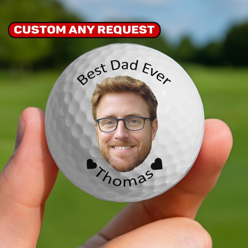 May include: A white golf ball with a personalized image of a man wearing glasses, with the text "Best Dad Ever" and the name "Thomas". Two black heart symbols are also present. The ball has the text "CUSTOM ANY REQUEST" in a red banner.