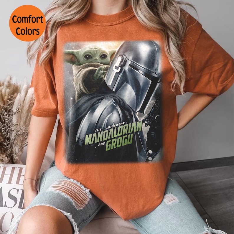 May include: Burnt orange t-shirt featuring a graphic from The Mandalorian series. The design includes the Mandalorian's helmet, Grogu, and the text "The Mandalorian and Grogu". The shirt has the words "Comfort Colors" in a circle.