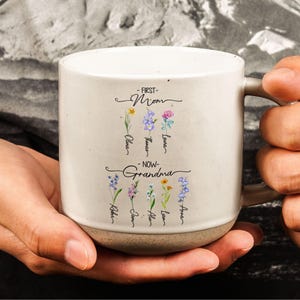 May include: A cream ceramic mug with a brown base, featuring floral illustrations and the text "-FIRST- Mom" and "-NOW- Grandma" with names beneath. The mug is held in a person's hands.