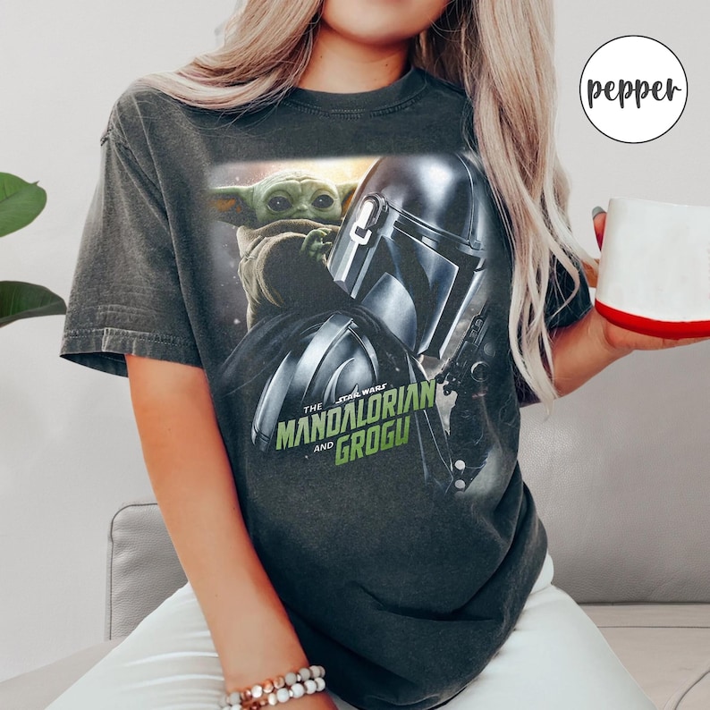May include: Dark gray t-shirt featuring a graphic of Grogu and the Mandalorian helmet from The Star Wars series. The text "The Mandalorian and Grogu" is printed in green and white. The shirt has short sleeves and a crew neck.