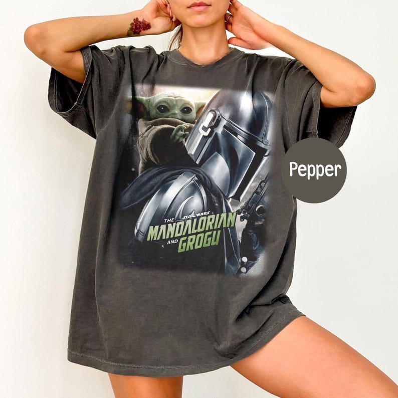 May include: A dark gray t-shirt featuring a graphic from The Mandalorian series. The design includes the Mandalorian's helmet, Grogu, and the text "The Mandalorian and Grogu" in green and white.