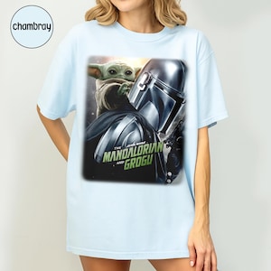 May include: Light blue t-shirt featuring a graphic of The Mandalorian and Grogu from Star Wars. The design includes the Mandalorian's helmet and Grogu, with the text "The Star Wars Mandalorian and Grogu".