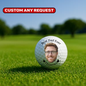 May include: A white golf ball with the text "Best Dad Ever" and a photo of a man wearing glasses. The name "Thomas" is below the photo, with two small black hearts. The ball is on green grass.