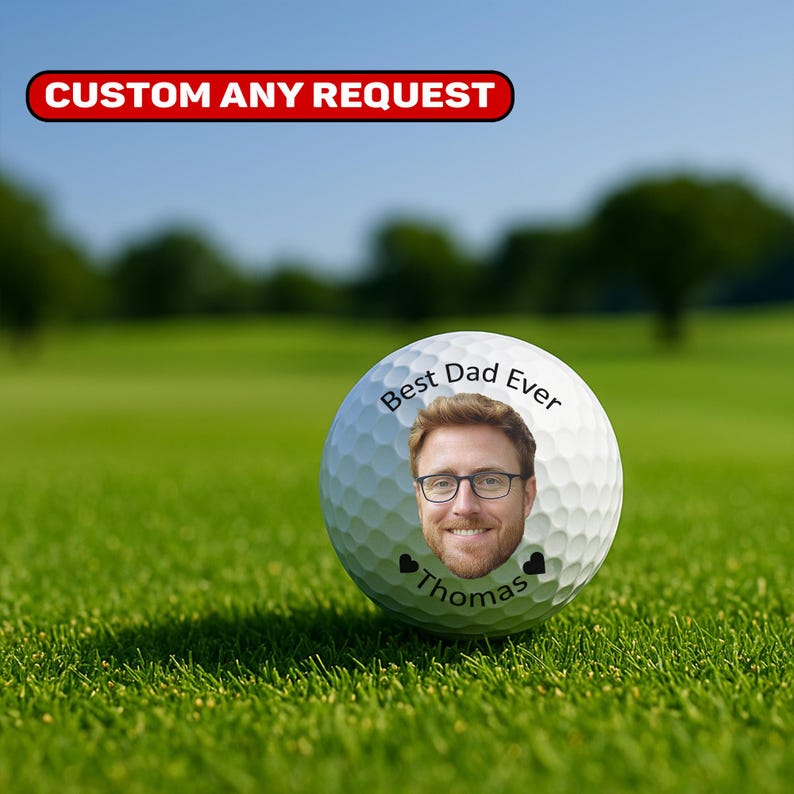 May include: A white golf ball with the text "Best Dad Ever" and a photo of a man wearing glasses. The name "Thomas" is below the photo, with two small black hearts. The ball is on green grass.
