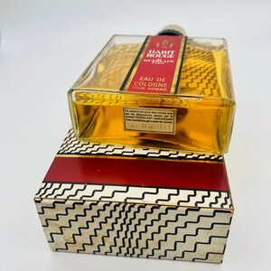 May include: A clear glass bottle of cologne with a gold label that reads "Habit Rouge Guerlain Eau de Cologne Pour Homme". The bottle is sitting on a red and white patterned box.