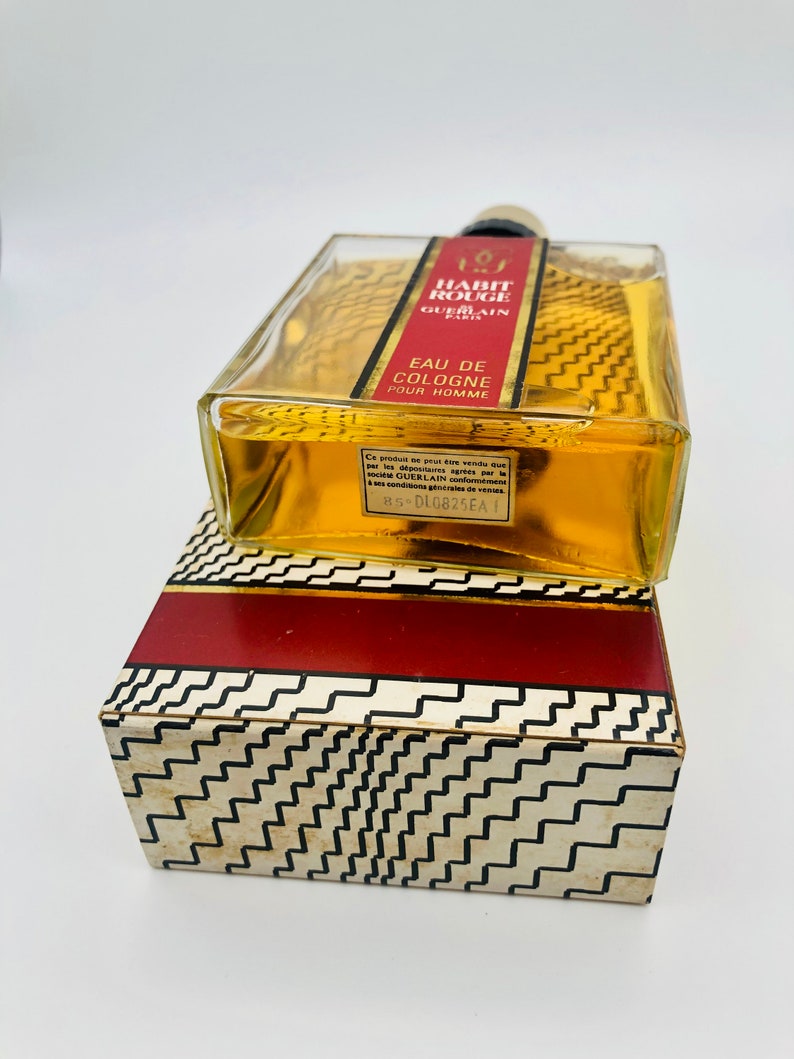 May include: A clear glass bottle of cologne with a gold label that reads "Habit Rouge Guerlain Eau de Cologne Pour Homme". The bottle is sitting on a red and white patterned box.