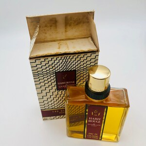 May include: A vintage bottle of Guerlain Habit Rouge cologne for men. The bottle is clear glass with a gold cap and a red label with the text "Habit Rouge Guerlain Paris" and "Eau de Cologne pour homme".