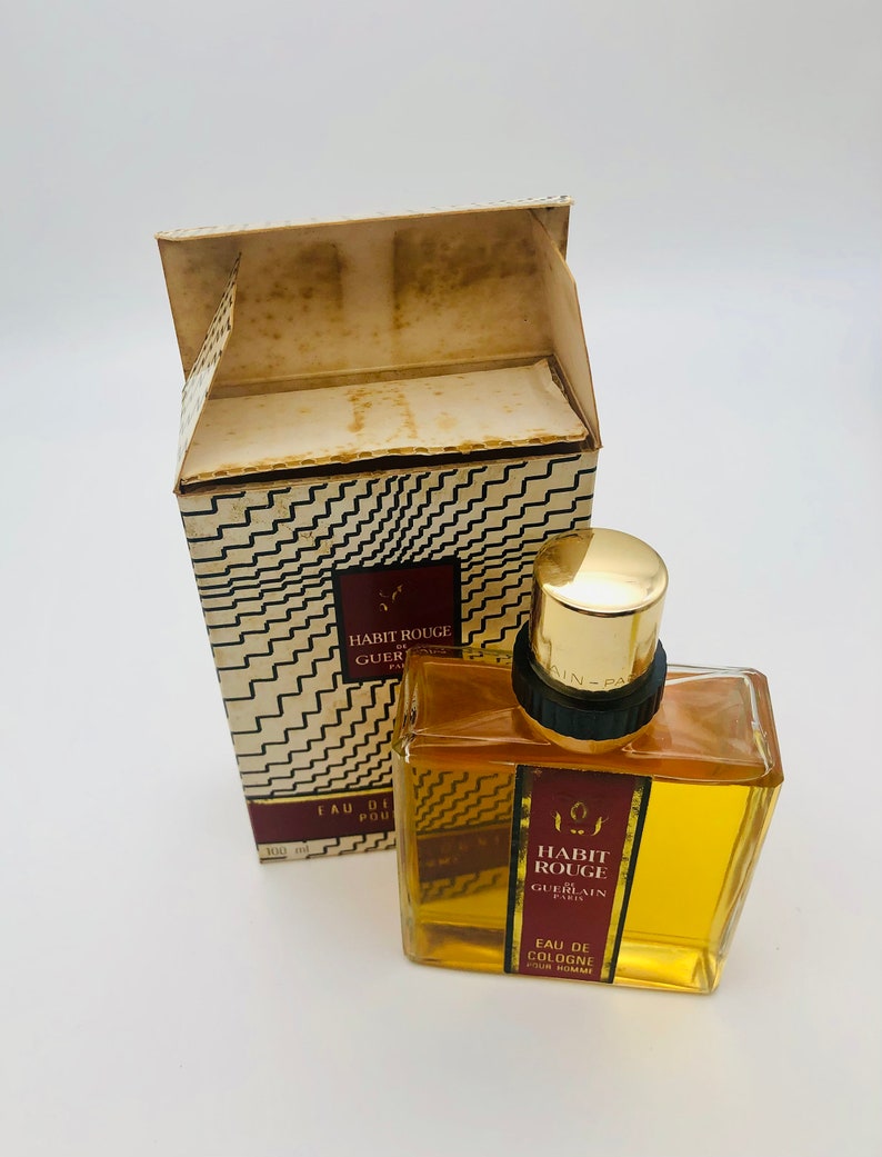 May include: A vintage bottle of Guerlain Habit Rouge cologne for men. The bottle is clear glass with a gold cap and a red label with the text "Habit Rouge Guerlain Paris" and "Eau de Cologne pour homme".