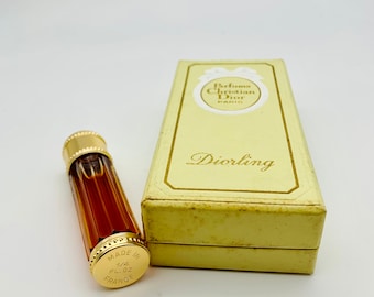 May include: A small, clear glass bottle of Diorling perfume with a gold cap and a gold label that says "Made in France 1/4 Fl. Oz.". The bottle is sitting on a white surface next to a white box with a gold label that says "Parfums Christian Dior Paris" and "Diorling".